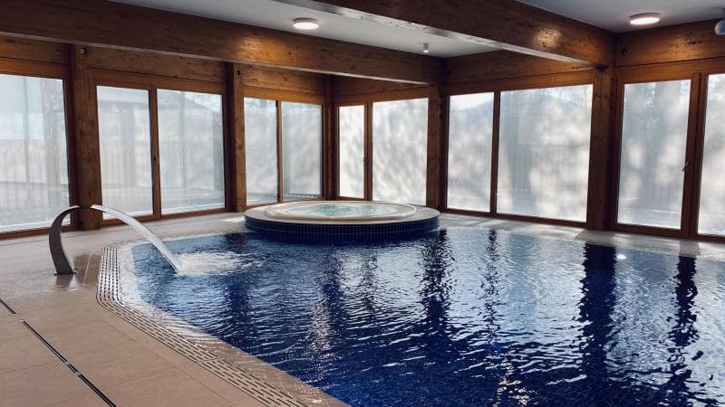 Überdachtes Schwimmbad mit Whirlpool, umgeben von Holzverkleidung und großen Fenstern.